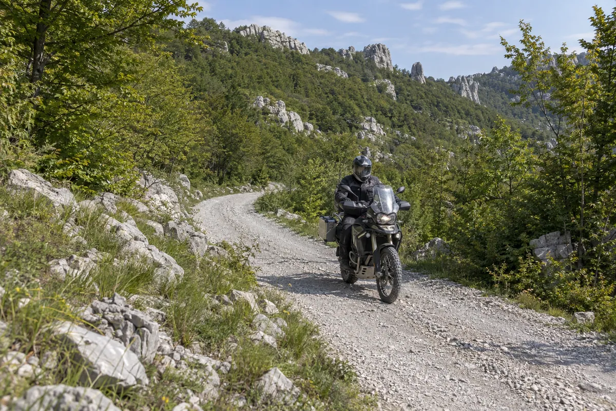 A rider follows pale gravel through green forest and the limestone forms of the Dabarski kukovi.