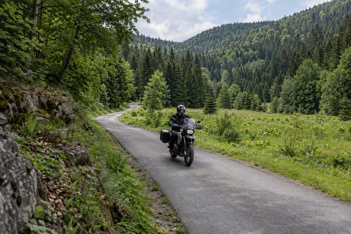 A motorcycle follows a narrow paved road through the conifer forest and green valley of Štirovača.
