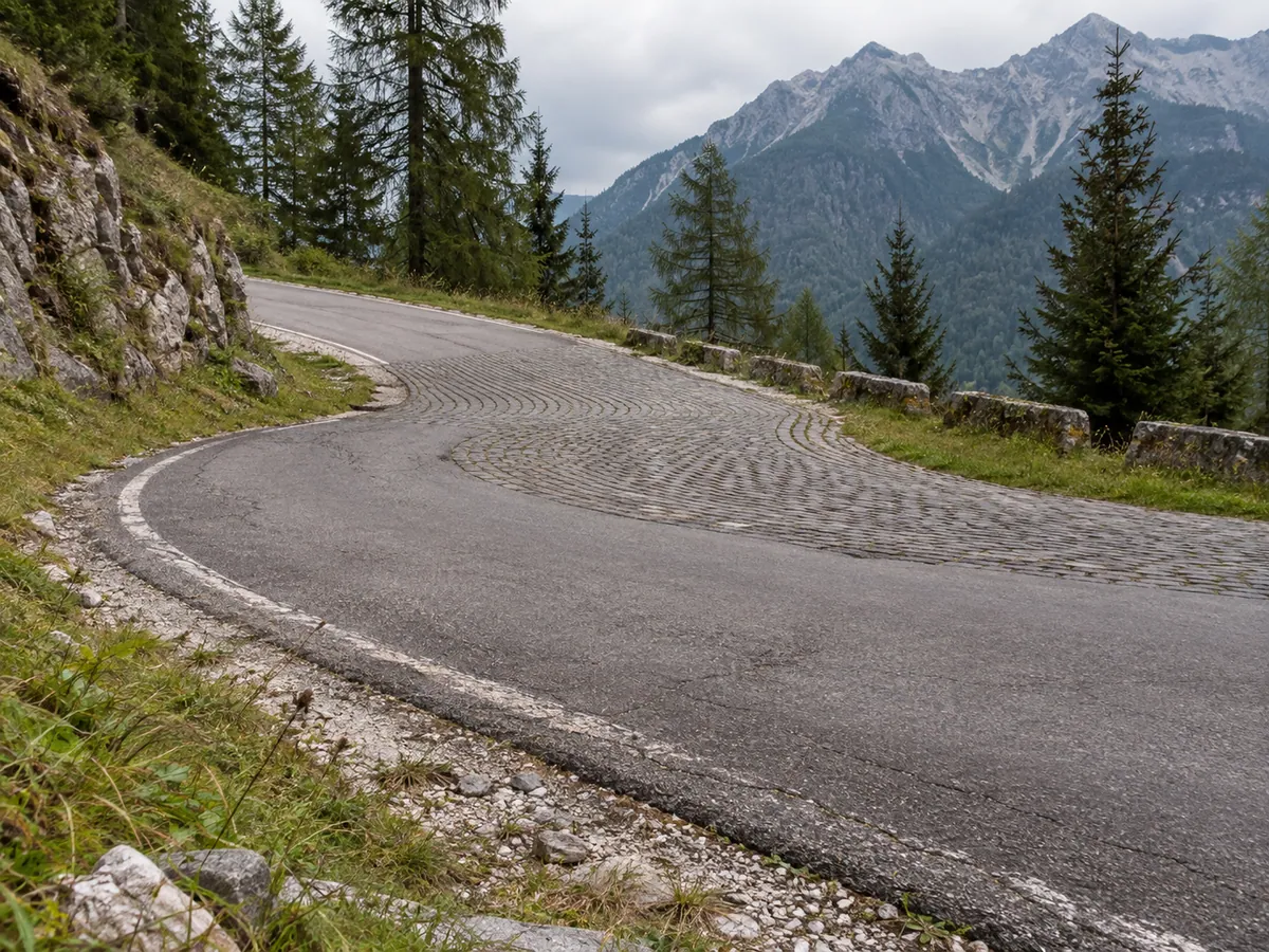 Asphalt meets stone-sett paving at a narrow mountain hairpin beside alpine vegetation.
